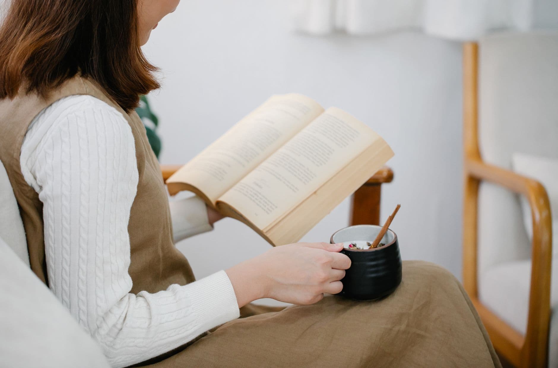 A woman reading in an armchair with a book in one hand and a cup of tea in the other
